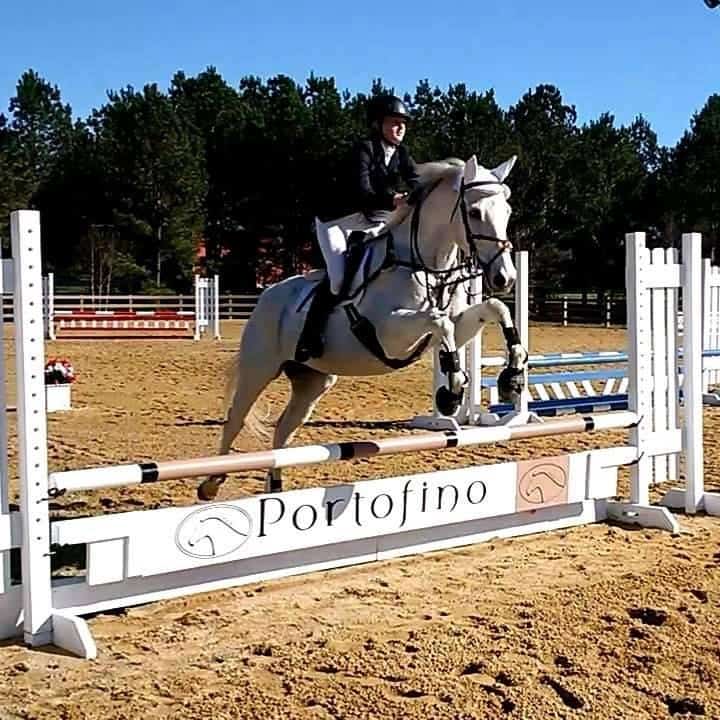 Rider on a white horse jumping in an arena outside.
