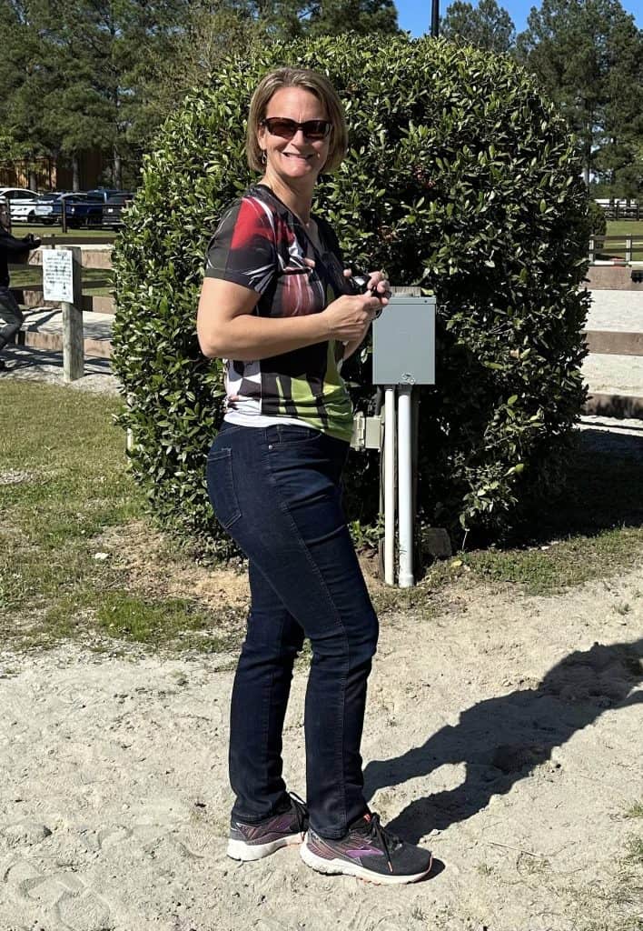 Smiling blond lady wearing sunglasses standing outside next to horse arena.