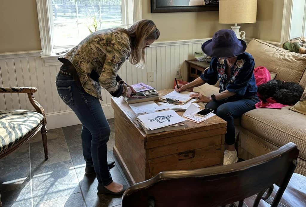 Woman sitting on couch writing on clipboard while another woman checks cellphone.